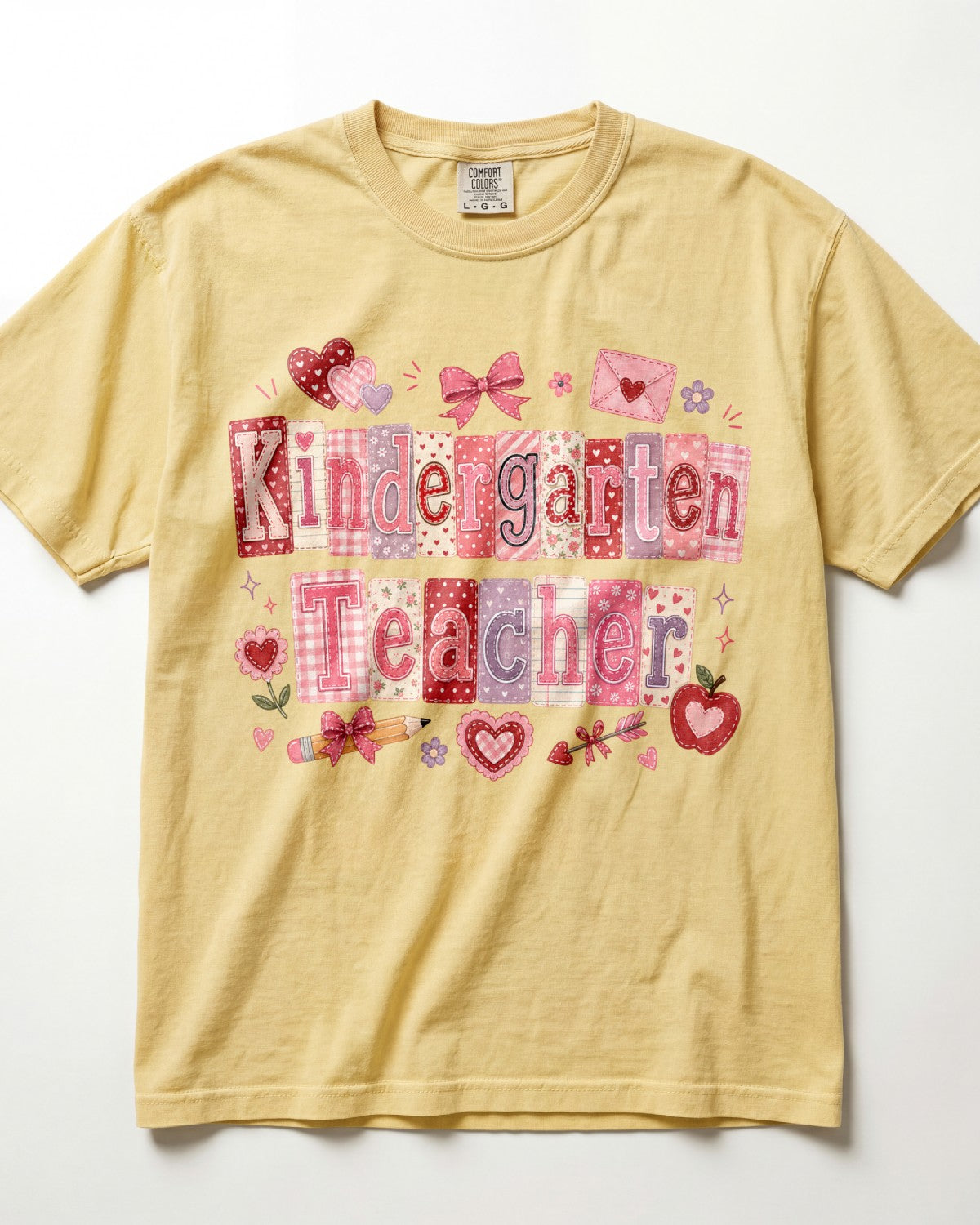 A pale yellow/butter short-sleeve t-shirt with a 'Kindergarten Teacher' graphic print in pink and red patchwork lettering, decorated with hearts, bows, and apples on a white background.