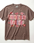 A brown/espresso short-sleeve crewneck t-shirt featuring a 'Kindergarten Teacher' graphic made of pink and red patterned letter tiles with hearts, bows, and envelopes, laid flat against a white background.