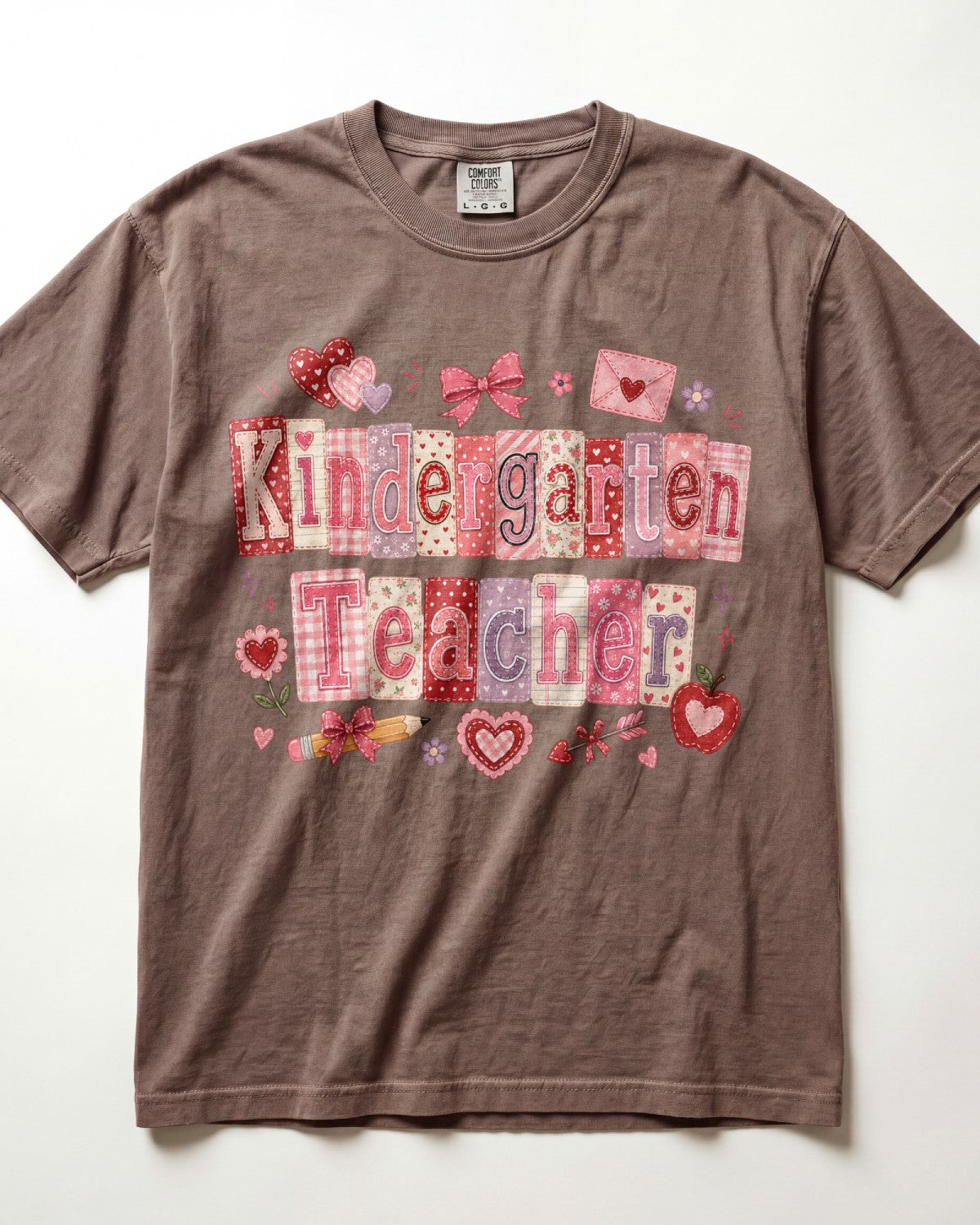 A brown/espresso short-sleeve crewneck t-shirt featuring a 'Kindergarten Teacher' graphic made of pink and red patterned letter tiles with hearts, bows, and envelopes, laid flat against a white background.