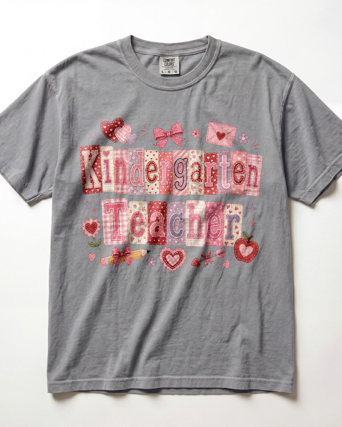 A gray short-sleeve crewneck t-shirt featuring a 'Kindergarten Teacher' graphic made of patchwork-style letters in pink, red, and white, decorated with hearts and bows.