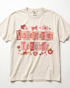 A cream-colored/ivory short-sleeve t-shirt featuring a 'Kindergarten Teacher' graphic print in pink, red, and beige patchwork patterns with school-themed illustrations like hearts, bows, and an apple.