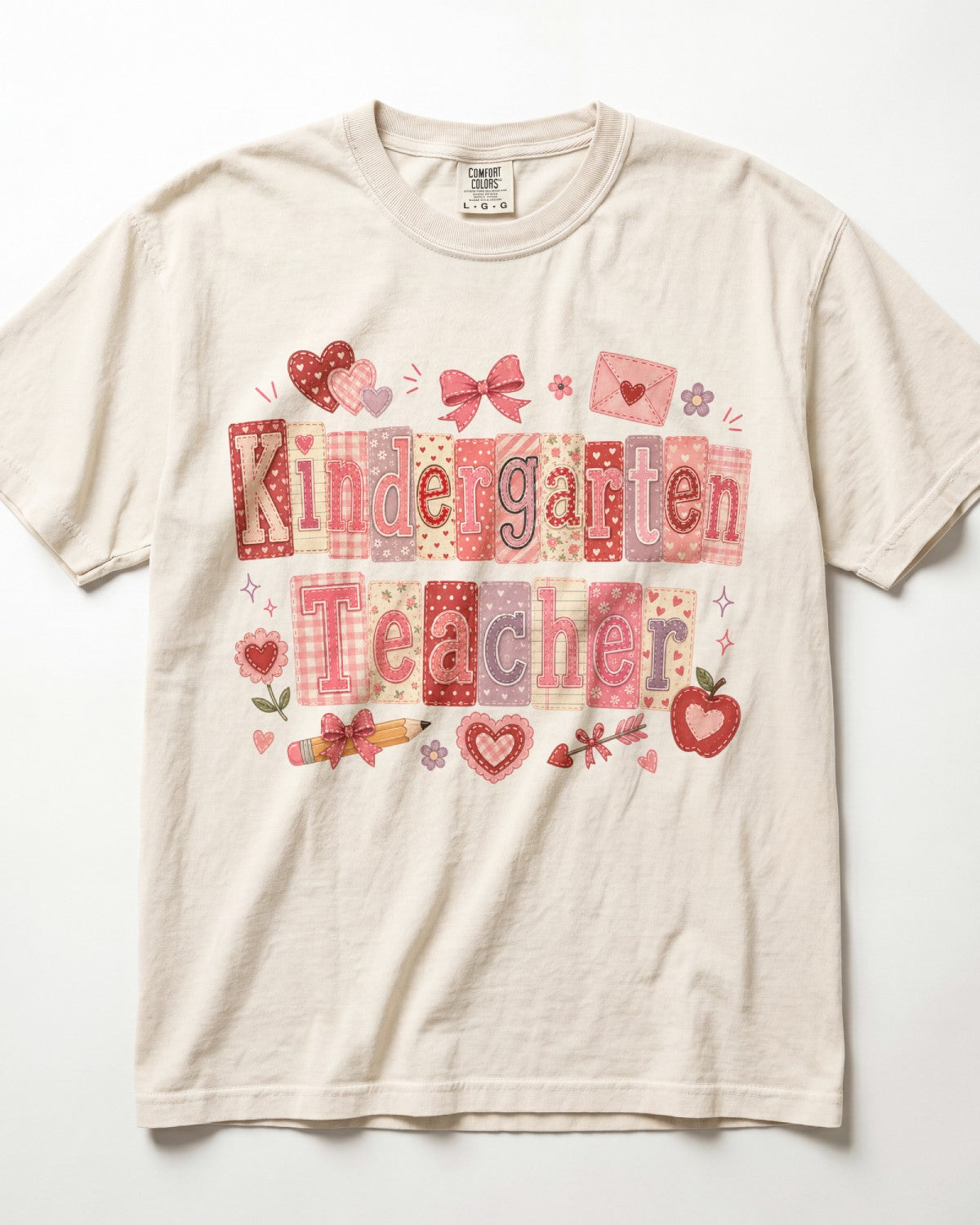 A cream-colored/ivory short-sleeve t-shirt featuring a 'Kindergarten Teacher' graphic print in pink, red, and beige patchwork patterns with school-themed illustrations like hearts, bows, and an apple.
