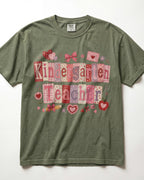 An olive green/moss short-sleeve t-shirt with a 'Kindergarten Teacher' patchwork graphic in pink, red, and purple tones, laid flat against a white background.