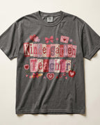 A dark gray/pepper crewneck t-shirt featuring a 'Kindergarten Teacher' graphic made of patchwork-style letters in pink, red, and white patterns, decorated with small hearts and bows.