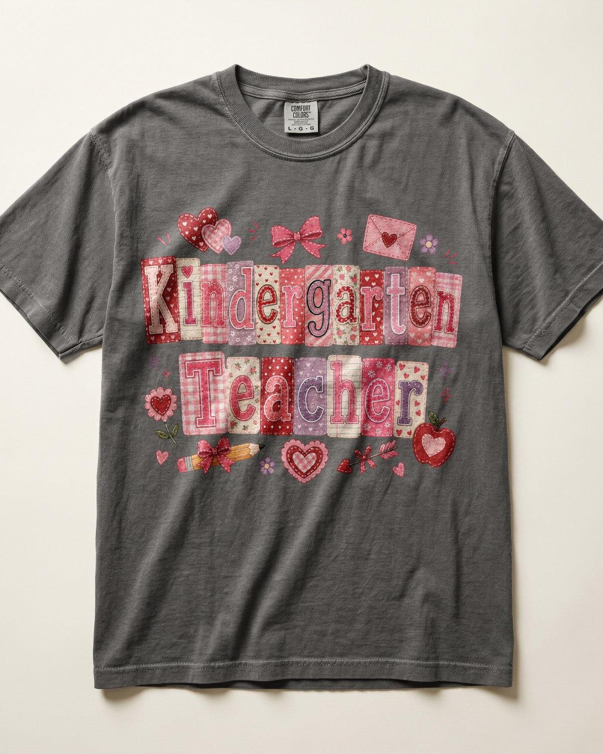 A dark gray/pepper crewneck t-shirt featuring a 'Kindergarten Teacher' graphic made of patchwork-style letters in pink, red, and white patterns, decorated with small hearts and bows.
