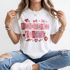 A woman wearing a white graphic t-shirt with a colorful 'Kindergarten Teacher' patchwork design, paired with distressed blue jeans and white sneakers.