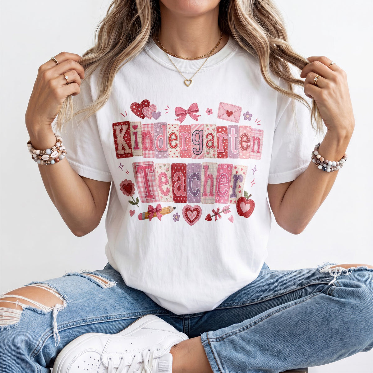 A woman wearing a white graphic t-shirt with a colorful 'Kindergarten Teacher' patchwork design, paired with distressed blue jeans and white sneakers.