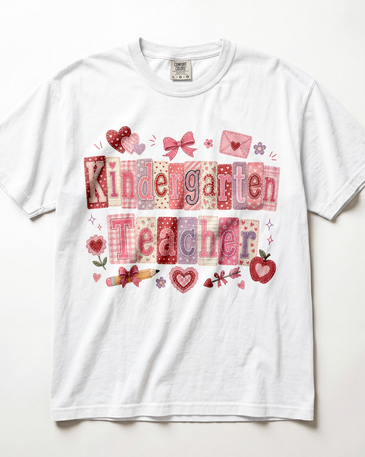 A white short-sleeve crewneck t-shirt featuring a 'Kindergarten Teacher' graphic made of pink and red patterned letter tiles, decorated with hearts, bows, and school-themed illustrations on a white background.